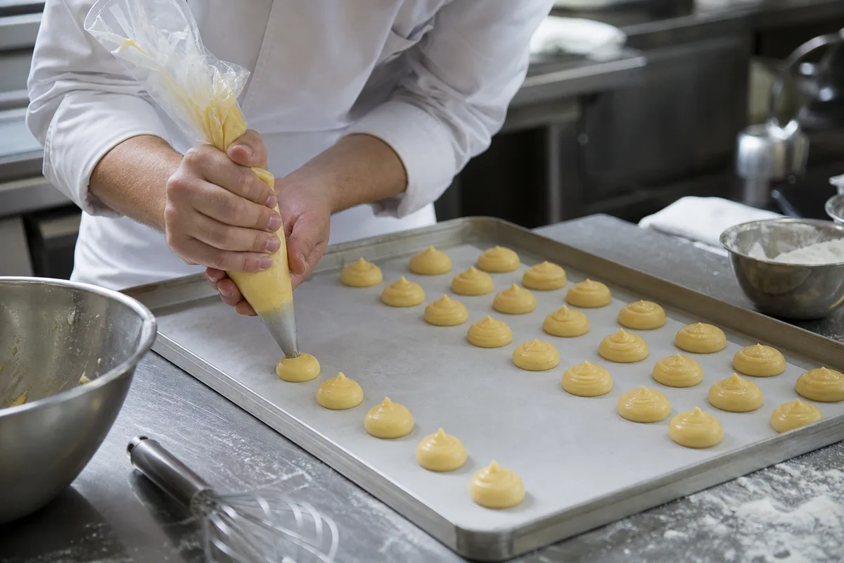 Apprenti en veste blanche dressant une poche à douille sur une plaque en laboratoire de pâtisserie